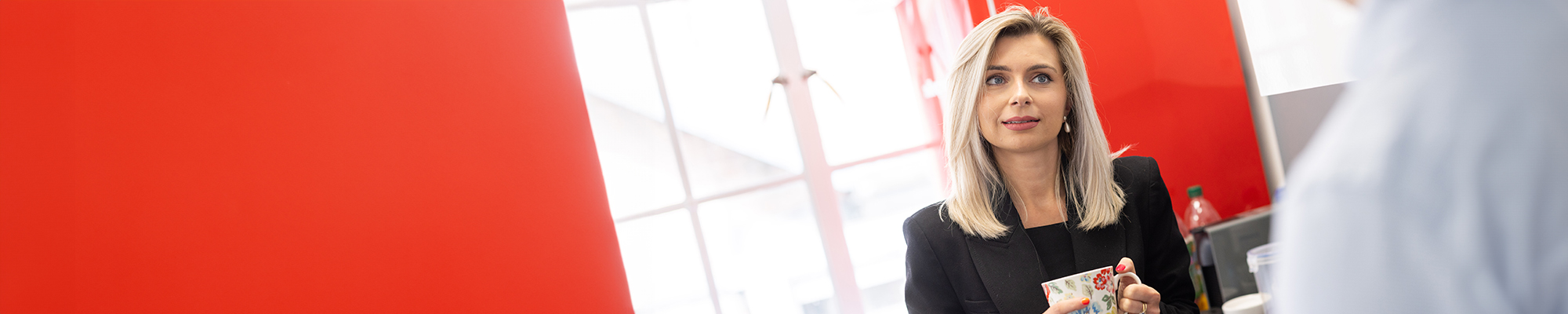 Lady with blonde hair having a serious conversation with another person, who has their back to us. The wall in the background is red and there are two windows in the room